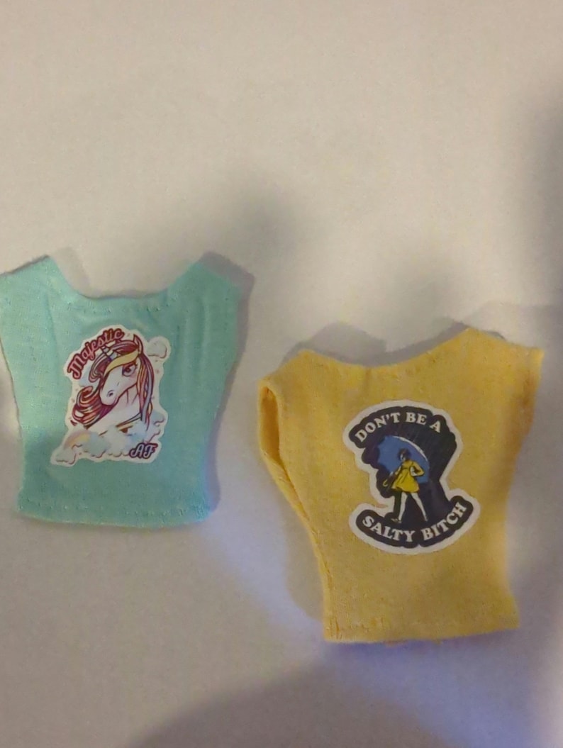 May include: Two doll-sized t-shirts. One is light blue with a unicorn graphic and the word "Majestic". The other is yellow with a graphic of a person under an umbrella and the text "Don't be a Salty Bitch".