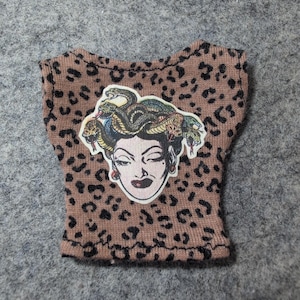 May include: A small, brown top with a leopard print pattern. The top features a Medusa graphic on the front. The Medusa graphic is a sticker with a white background and a detailed illustration of Medusa's face and snakes.