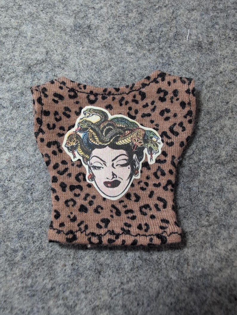 May include: A small, brown top with a leopard print pattern. The top features a Medusa graphic on the front. The Medusa graphic is a sticker with a white background and a detailed illustration of Medusa's face and snakes.