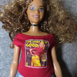 May include: A doll with brown curly hair, blue eyes, and freckles, wearing a red off-the-shoulder shirt with the text "SHE TRIED TO BE GOOD" and a graphic of a woman at a bus stop. The doll is also wearing a black choker and blue jeans.