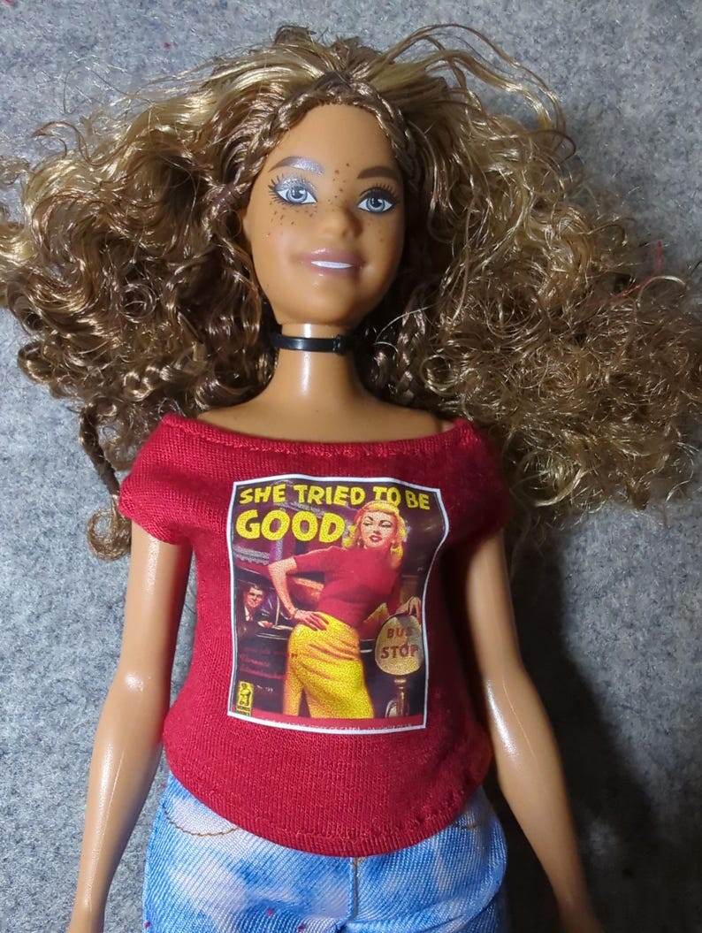 May include: A doll with brown curly hair, blue eyes, and freckles, wearing a red off-the-shoulder shirt with the text "SHE TRIED TO BE GOOD" and a graphic of a woman at a bus stop. The doll is also wearing a black choker and blue jeans.