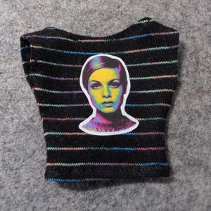 May include: Black crop top with horizontal rainbow stripes. A colorful portrait of a woman is centered on the front. The top is made of a soft, knit fabric. The overall theme is fashion.