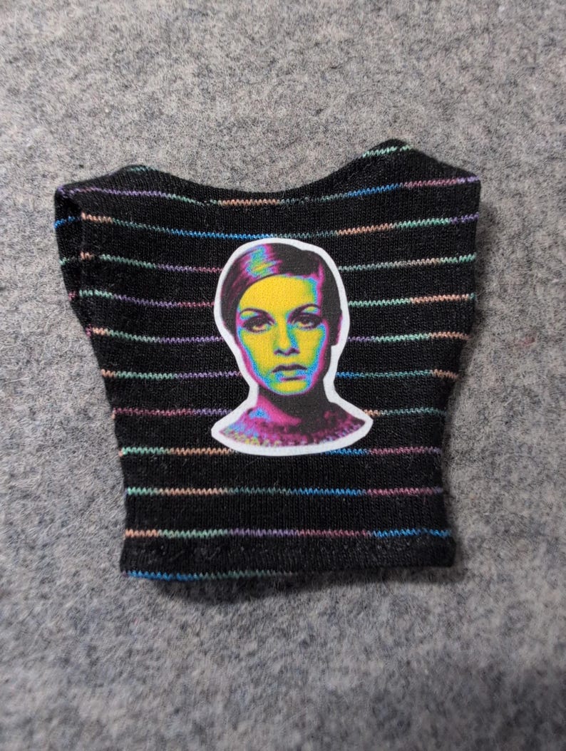 May include: Black crop top with horizontal rainbow stripes. A colorful portrait of a woman is centered on the front. The top is made of a soft, knit fabric. The overall theme is fashion.