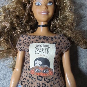 May include: A doll with brown skin, blue eyes, and curly brown hair. The doll wears a black choker, a brown leopard print top with the text "Josephine Baker", and blue jeans. The top features an illustration of Josephine Baker.