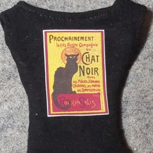May include: Black miniature shirt featuring a vintage-style poster for Le Chat Noir. The poster has a black cat silhouette, text in yellow and red, and the words "PROCHAINEMENT" and "CHAT NOIR".