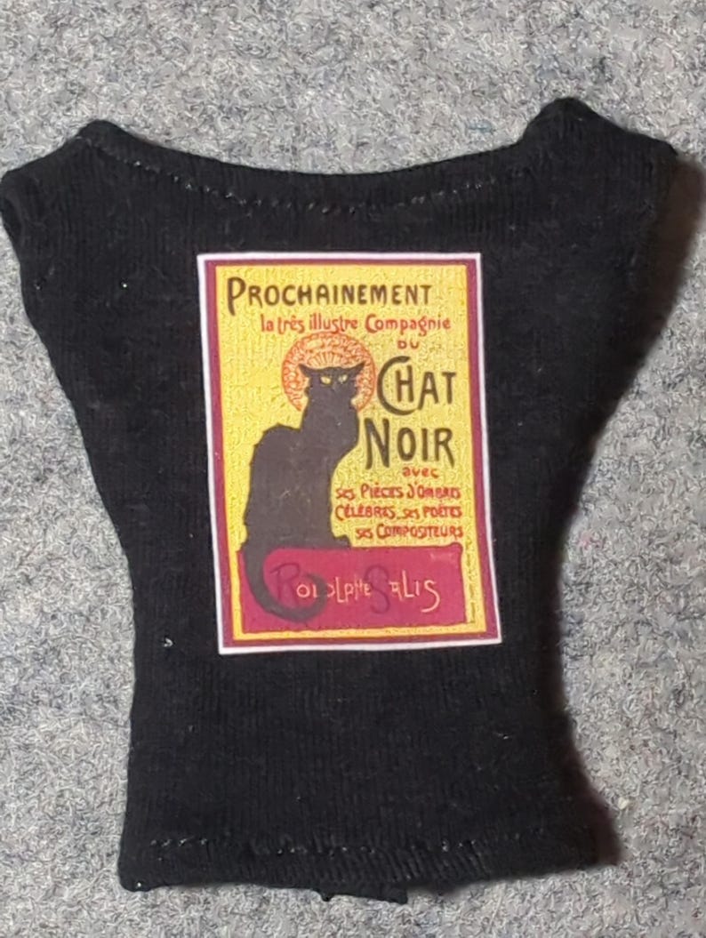 May include: Black miniature shirt featuring a vintage-style poster for Le Chat Noir. The poster has a black cat silhouette, text in yellow and red, and the words "PROCHAINEMENT" and "CHAT NOIR".