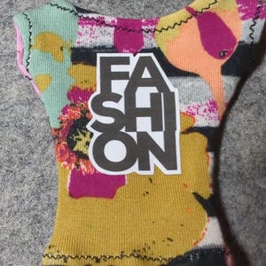 May include: A colorful doll-sized top with the word "FASHION" in bold black letters. The top features a vibrant mix of colors, including yellow, pink, orange, and green, with a textured fabric and black stitching.