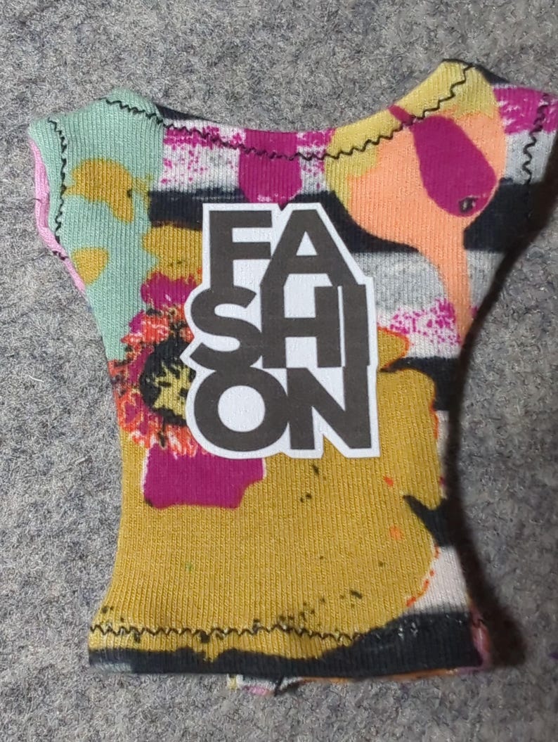 May include: A colorful doll-sized top with the word "FASHION" in bold black letters. The top features a vibrant mix of colors, including yellow, pink, orange, and green, with a textured fabric and black stitching.