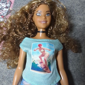 May include: A doll with curly brown hair, blue eyes, and freckles, wearing a light blue t-shirt with a vintage surfing graphic and denim shorts. The t-shirt features the name "Hilda" and a surfer girl illustration. A black choker necklace is around the doll's neck.