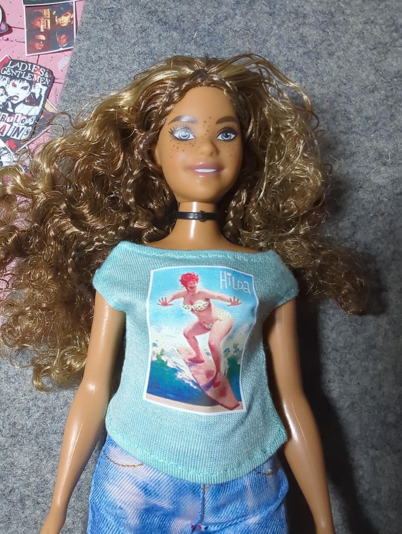 May include: A doll with curly brown hair, blue eyes, and freckles, wearing a light blue t-shirt with a vintage surfing graphic and denim shorts. The t-shirt features the name "Hilda" and a surfer girl illustration. A black choker necklace is around the doll's neck.