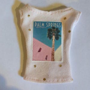 May include: A light pink t-shirt with a Palm Springs graphic. The graphic features a palm tree and the words "Palm Springs" against a backdrop of pink, blue, and yellow. The shirt has gold embellishments.