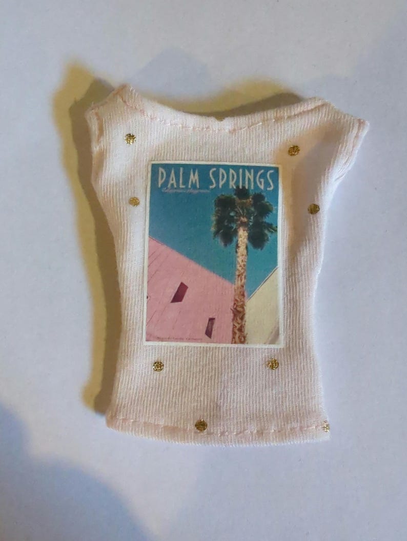 May include: A light pink t-shirt with a Palm Springs graphic. The graphic features a palm tree and the words "Palm Springs" against a backdrop of pink, blue, and yellow. The shirt has gold embellishments.