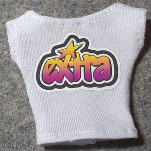 May include: White sleeveless top with the word "EXTRA" in a gradient of yellow, orange, and purple, with a yellow star graphic. The text is outlined in black. The top is made of a ribbed fabric.