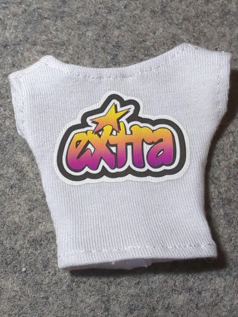 May include: White sleeveless top with the word "EXTRA" in a gradient of yellow, orange, and purple, with a yellow star graphic. The text is outlined in black. The top is made of a ribbed fabric.