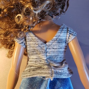 May include: A doll wearing a gray and white knit top and blue denim jeans. The top has a V-neck and short sleeves. The doll has long, curly blonde hair.
