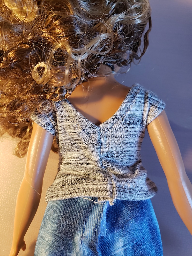 May include: A doll wearing a gray and white knit top and blue denim jeans. The top has a V-neck and short sleeves. The doll has long, curly blonde hair.