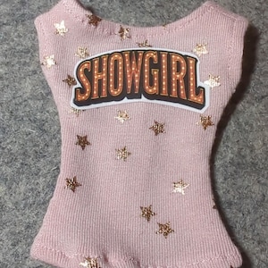 May include: A light pink tank top with gold star accents and the word "SHOWGIRL" in a marquee-style design. The top is made of a ribbed fabric.