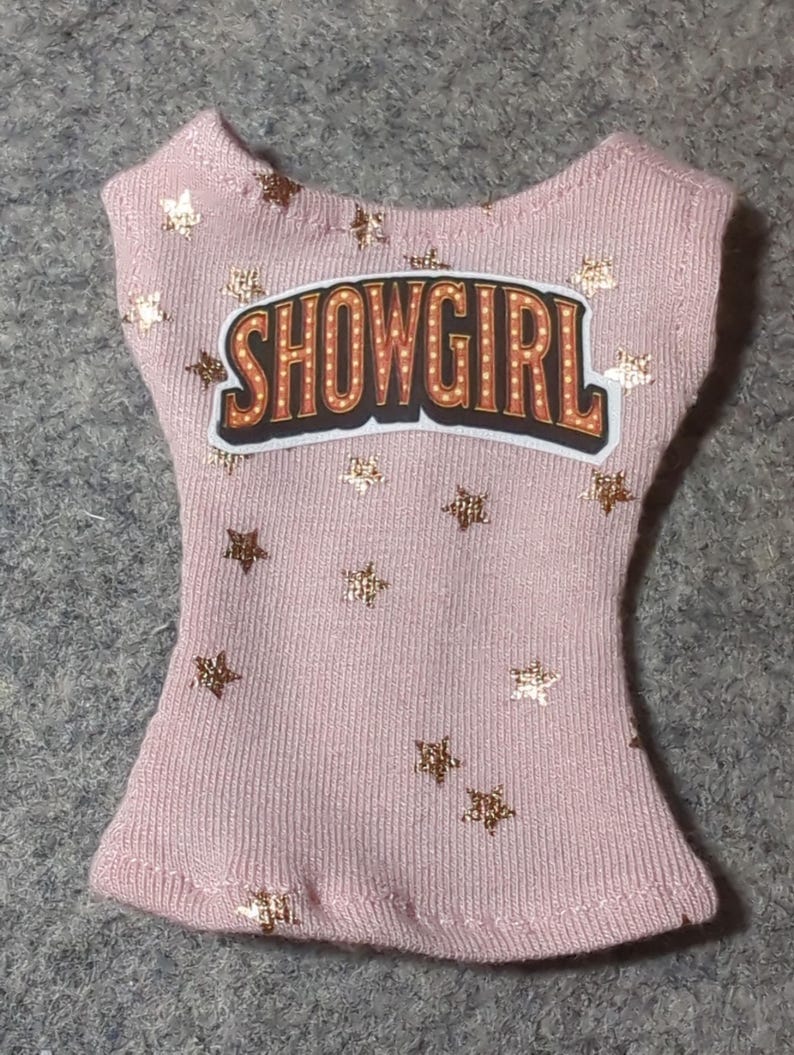 May include: A light pink tank top with gold star accents and the word "SHOWGIRL" in a marquee-style design. The top is made of a ribbed fabric.