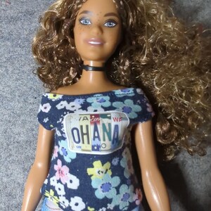 May include: A doll with tan skin, blue eyes, and curly blonde hair. She wears a dark blue off-the-shoulder top with a floral pattern and the word "OHANA" on the front. She also wears a black choker and blue denim shorts.