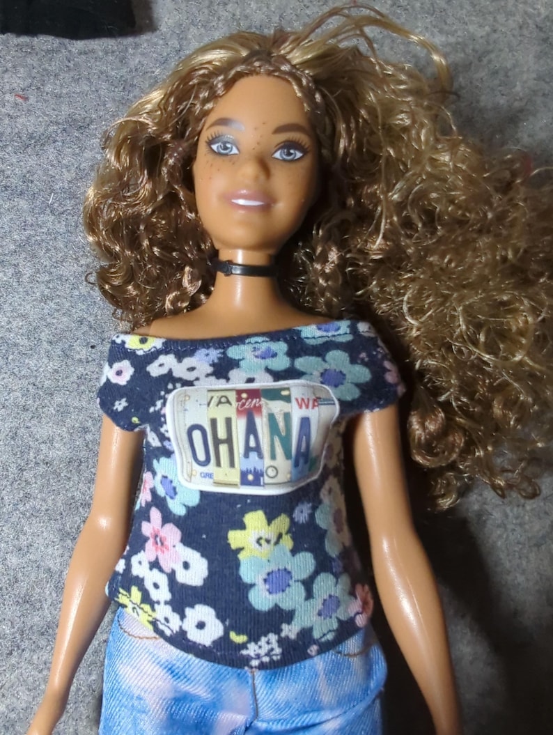 May include: A doll with tan skin, blue eyes, and curly blonde hair. She wears a dark blue off-the-shoulder top with a floral pattern and the word "OHANA" on the front. She also wears a black choker and blue denim shorts.