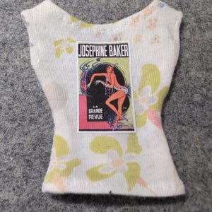 May include: A white sleeveless top with a floral pattern in yellow, pink, and orange. The top features a rectangular graphic of Josephine Baker with the text "LA GRANDE REVUE".