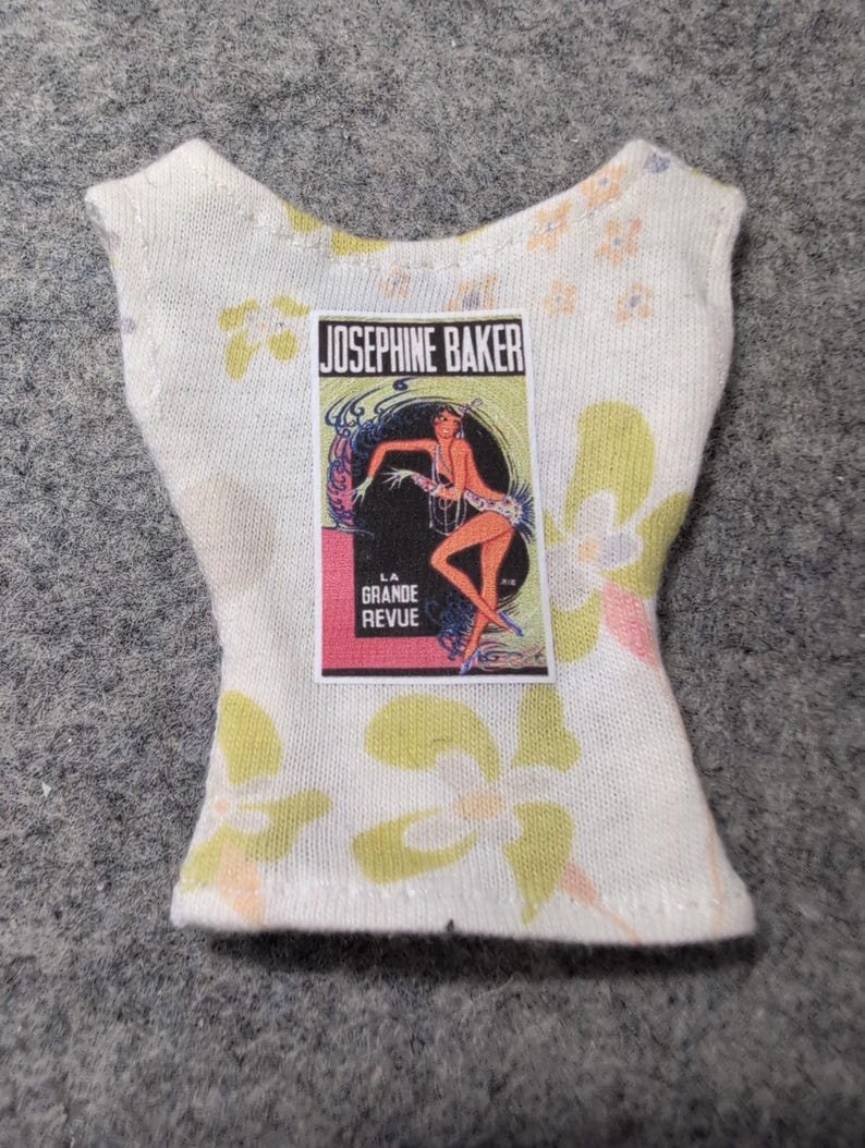 May include: A white sleeveless top with a floral pattern in yellow, pink, and orange. The top features a rectangular graphic of Josephine Baker with the text "LA GRANDE REVUE".