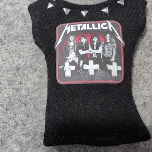 May include: Black doll-sized t-shirt with the band name "METALLICA" printed on the front. The graphic features the band members and crosses. Silver-colored studs are placed along the neckline.