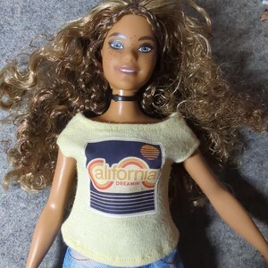 May include: A doll with curly brown hair, blue eyes, and freckles, wearing a light yellow off-the-shoulder top with a "California Dreamin'" graphic and blue tie-dye jeans. The doll has a black choker necklace.