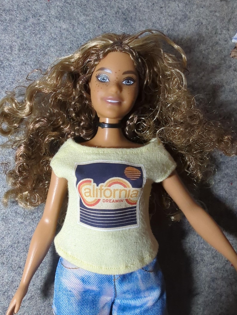 May include: A doll with curly brown hair, blue eyes, and freckles, wearing a light yellow off-the-shoulder top with a "California Dreamin'" graphic and blue tie-dye jeans. The doll has a black choker necklace.