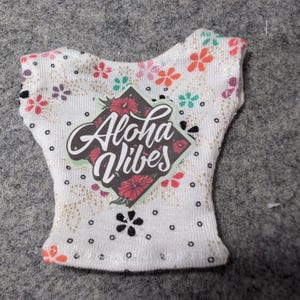 May include: White sleeveless top with a floral pattern and the words "Aloha Vibes" in a diamond-shaped graphic. The top features a variety of colorful flowers and black polka dots. The fabric has a subtle glittery sheen.