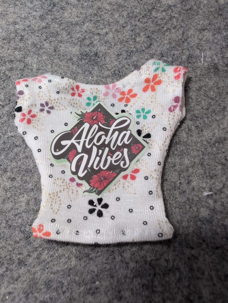 May include: White sleeveless top with a floral pattern and the words "Aloha Vibes" in a diamond-shaped graphic. The top features a variety of colorful flowers and black polka dots. The fabric has a subtle glittery sheen.