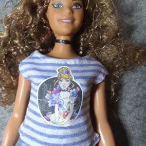 May include: A doll with curly brown hair, blue eyes, and freckles. The doll wears a striped blue and white t-shirt with a graphic of a princess, a black choker, and blue tie-dye jeans. The doll's skin is tan.