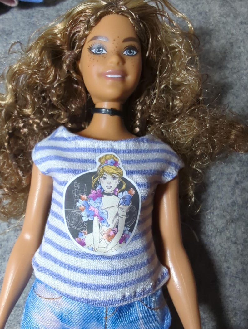 May include: A doll with curly brown hair, blue eyes, and freckles. The doll wears a striped blue and white t-shirt with a graphic of a princess, a black choker, and blue tie-dye jeans. The doll's skin is tan.