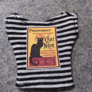 May include: A black and white striped top featuring a vintage "Chat Noir" poster design. The poster includes text and a black cat silhouette on a yellow background. The top is a doll-sized garment.