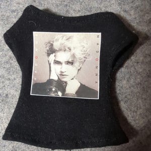 May include: Black doll-sized t-shirt featuring a black and white image of Madonna. The image is framed with the word "MADONNA" printed vertically on both sides. The shirt has a scoop neck and short sleeves.
