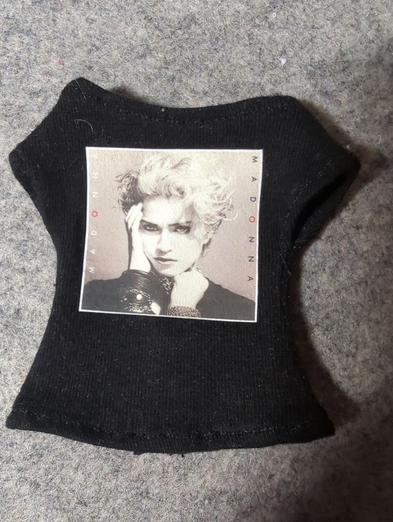May include: Black doll-sized t-shirt featuring a black and white image of Madonna. The image is framed with the word "MADONNA" printed vertically on both sides. The shirt has a scoop neck and short sleeves.