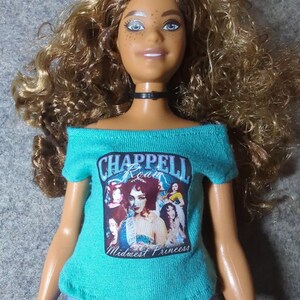 May include: A doll with long, curly blonde hair and blue eyes wearing a teal off-the-shoulder shirt with the text "Chappell Roan Midwest Princess" and blue and white patterned shorts. The doll has a black choker.