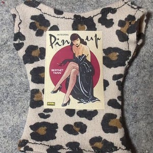 May include: A beige and brown leopard print top with a pin-up graphic. The graphic features a woman in a black dress and the text "INTEGRAL Pin-up" and "BERTHET YANN". The top has a scoop neck and cap sleeves.