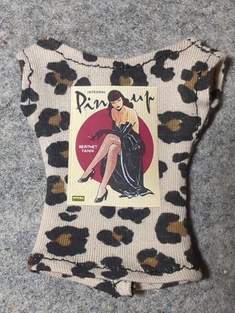 May include: A beige and brown leopard print top with a pin-up graphic. The graphic features a woman in a black dress and the text "INTEGRAL Pin-up" and "BERTHET YANN". The top has a scoop neck and cap sleeves.
