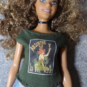 May include: A doll with curly blonde hair, blue eyes, and freckles, wearing a black choker and a green t-shirt. The t-shirt features a vintage-style graphic with the words "ABSINTHE la Fee Verte."