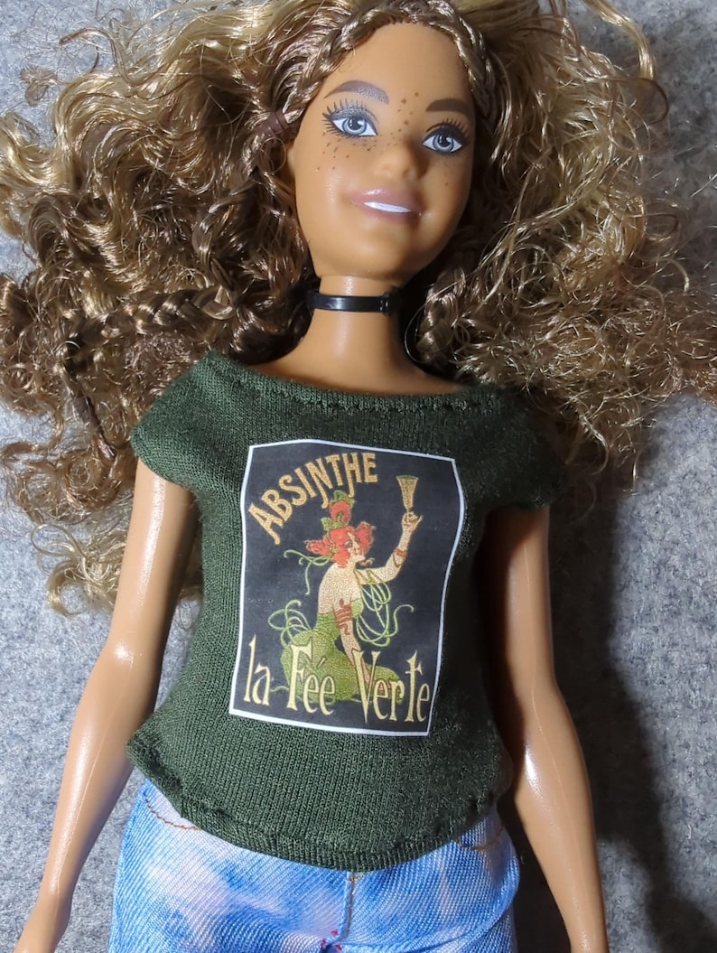 May include: A doll with curly blonde hair, blue eyes, and freckles, wearing a black choker and a green t-shirt. The t-shirt features a vintage-style graphic with the words "ABSINTHE la Fee Verte."
