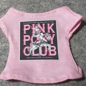 May include: A light pink doll-sized t-shirt with a graphic that reads "PINK PONY CLUB" with a carousel horse design. The text "I'M GONNA KEEP ON DANCING AT THE" and "WEST HOLLYWOOD, CALIFORNIA" are also printed on the shirt.