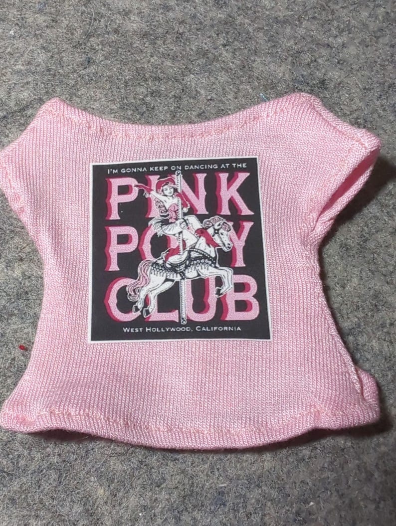 May include: A light pink doll-sized t-shirt with a graphic that reads "PINK PONY CLUB" with a carousel horse design. The text "I'M GONNA KEEP ON DANCING AT THE" and "WEST HOLLYWOOD, CALIFORNIA" are also printed on the shirt.