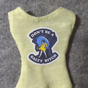 May include: A light yellow doll-sized top with a graphic that reads "DON'T BE A SALTY BITCH." The graphic features a cartoon person under a blue umbrella in the rain. The top is made of a soft, textured fabric.