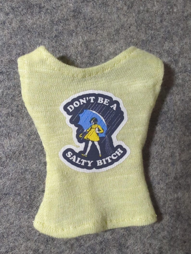 May include: A light yellow doll-sized top with a graphic that reads "DON'T BE A SALTY BITCH." The graphic features a cartoon person under a blue umbrella in the rain. The top is made of a soft, textured fabric.