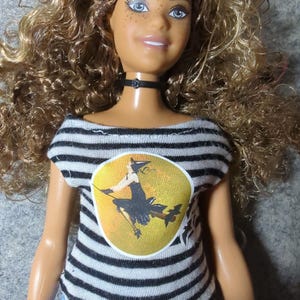 May include: A doll with curly blonde hair, blue eyes, and freckles, wearing a black choker and a black and white striped shirt. The shirt features a yellow circle with a witch graphic.