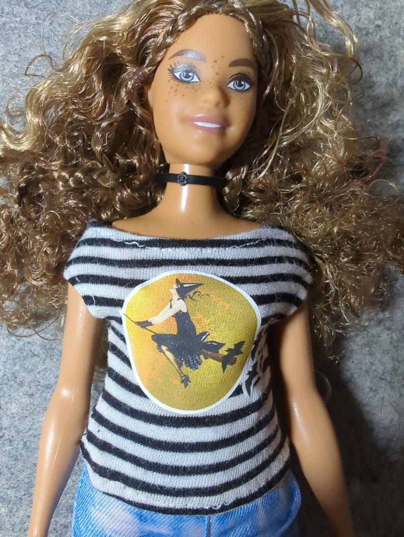 May include: A doll with curly blonde hair, blue eyes, and freckles, wearing a black choker and a black and white striped shirt. The shirt features a yellow circle with a witch graphic.