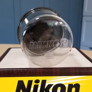 May include: A clear glass lens cap featuring the embossed text NIKKOR and a smaller NIPPON KOGAKU TOKYO logo. The circular cap is transparent with a smooth surface, displaying the brand name clearly in the center of the glass lens cover.