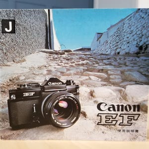 以下が含まれることがあります： 石畳と建物を背景にした、レンズ付きの黒いCanon EFカメラ。カメラにはCanonのロゴとEFのモデル名があります。画像には、左上隅に文字「J」も含まれています。