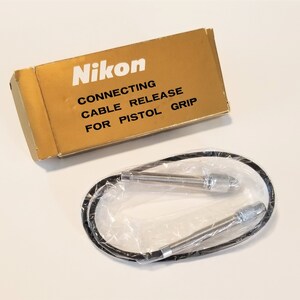 Può includere: Una scatola Nikon dorata con la scritta "Nikon" e "Connecting Cable Release for Pistol Grip." Un rilascio del cavo nero è in un sacchetto di plastica trasparente.